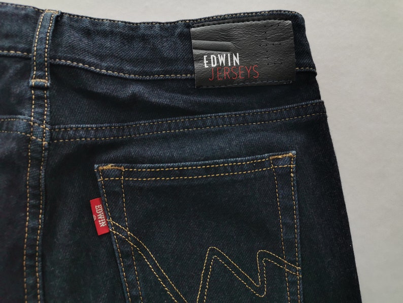 K&ouml;nnte beinhalten: Nahaufnahme einer dunkelblauen Jeans mit gelben N&auml;hten. Die Ges&auml;&szlig;tasche hat ein markantes 'W'-Nahtdesign. Ein rotes Edwin-Etikett ist auf der Tasche sichtbar, und ein schwarzes Lederpatch mit 'Edwin Jerseys' befindet sich am Bund.