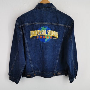 May include: A dark blue denim jacket with a Universal Studios Japan logo patch on the back. The patch features a globe design with the words "UNIVERSAL STUDIOS JAPAN" in yellow and red lettering.