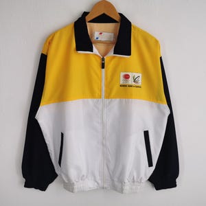 May include: A vintage yellow, white, and black zip-up jacket. The jacket features a black collar and sleeves, a yellow upper chest, and a white lower chest. The jacket has a small logo on the upper chest that reads "KEWIN 2000 in Sydney".
