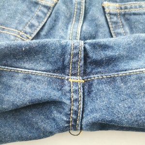 May include: Close-up of blue denim jeans. The image highlights the stitching details, with yellow thread creating seams and accents. The fabric has a textured appearance, and the overall tone is a classic, worn denim look.