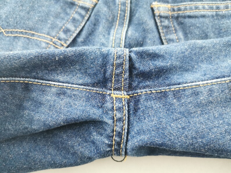 May include: Close-up of blue denim jeans. The image highlights the stitching details, with yellow thread creating seams and accents. The fabric has a textured appearance, and the overall tone is a classic, worn denim look.