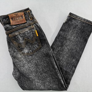 May include: Acid wash denim jeans with a faded black and gray pattern. Features a brown leather patch with the "EDWIN" logo, a yellow tag, and a back pocket with a yellow stitched "W" design.