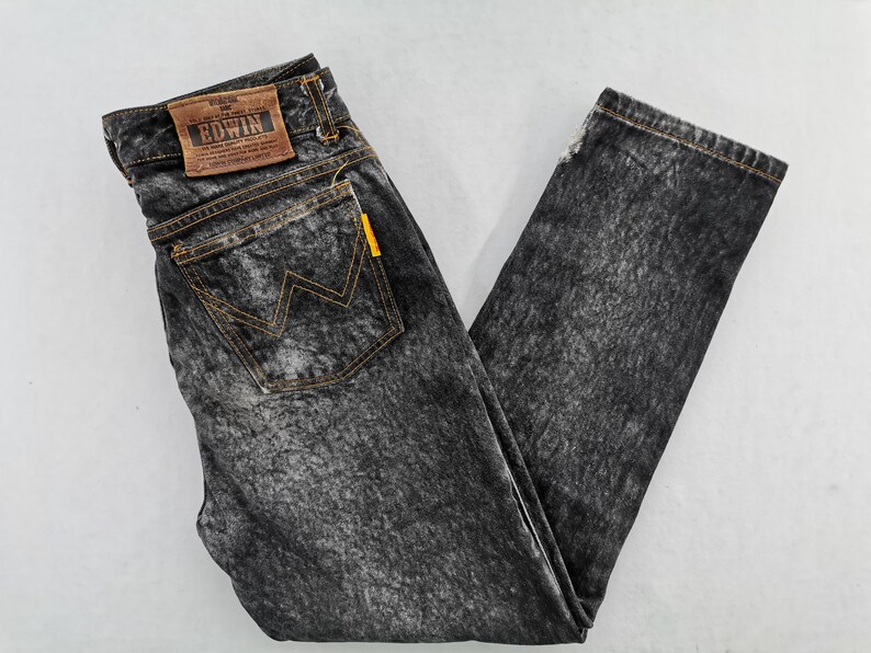 May include: Acid wash denim jeans with a faded black and gray pattern. Features a brown leather patch with the "EDWIN" logo, a yellow tag, and a back pocket with a yellow stitched "W" design.