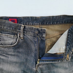 May include: Close-up of a pair of faded blue denim jeans with a partially unzipped fly. The jeans have a classic five-pocket design, a button closure, and a red tag with white text. The zipper is blue.