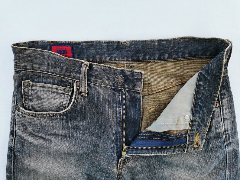 May include: Close-up of a pair of faded blue denim jeans with a partially unzipped fly. The jeans have a classic five-pocket design, a button closure, and a red tag with white text. The zipper is blue.
