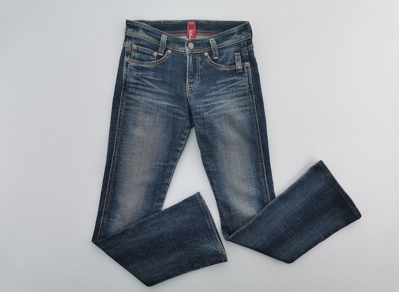Pu&ograve; includere: Jeans in denim blu scuro con lavaggio sbiadito, caratterizzati da un classico design a cinque tasche, chiusura con bottone e zip e una leggera svasatura sul fondo delle gambe. La vita presenta passanti per cintura e un'etichetta rossa interna.