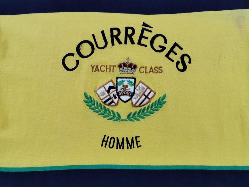 Op de afbeelding: Gele stof met het woord "COURR&Egrave;GES" in zwart, bovenaan gebogen. Daaronder staat "YACHT CLASS" in goud. Een wapen met een kroon en schilden is gecentreerd, met "SPORT" in het midden. Het woord "HOMME" staat onderaan.