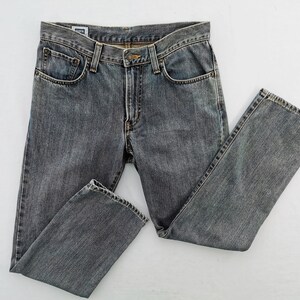 May include: Gray denim jeans with a classic five-pocket design. The jeans have a button and zipper closure, belt loops, and a faded appearance. The garment is laid flat on a white surface, showcasing the details of the fabric and stitching.