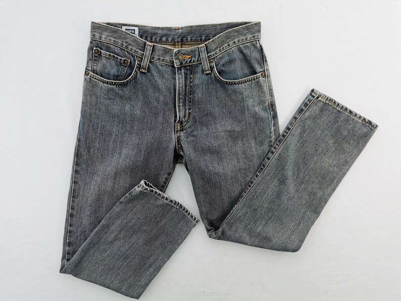 May include: Gray denim jeans with a classic five-pocket design. The jeans have a button and zipper closure, belt loops, and a faded appearance. The garment is laid flat on a white surface, showcasing the details of the fabric and stitching.