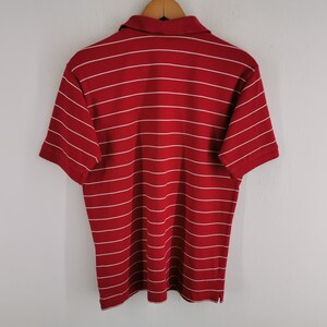 Puede incluir: Polo rojo con rayas horizontales blancas. La camiseta de manga corta est&aacute; colgada de una percha de madera. La camisa tiene cuello y tapeta abotonada.