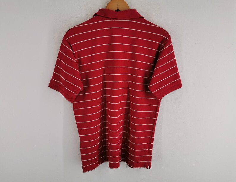 Puede incluir: Polo rojo con rayas horizontales blancas. La camiseta de manga corta est&aacute; colgada de una percha de madera. La camisa tiene cuello y tapeta abotonada.