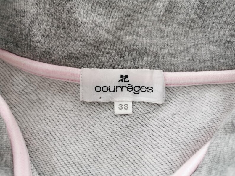May include: Close-up of a gray sweatshirt collar with a pink trim. A white label displays the brand name "courr&egrave;ges" and the size "38". The fabric has a textured appearance.