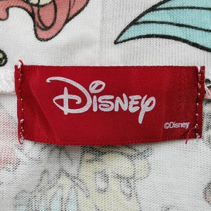 May include: Close-up of a red Disney label with the Disney logo in white. The label is sewn onto a white fabric with colorful cartoon characters. The fabric has a pattern of cartoon characters.