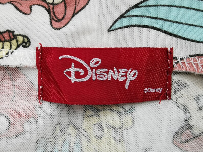 May include: Close-up of a red Disney label with the Disney logo in white. The label is sewn onto a white fabric with colorful cartoon characters. The fabric has a pattern of cartoon characters.
