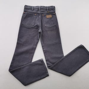 May include: Dark gray denim jeans with a classic five-pocket design. The jeans feature a brown leather patch and copper rivets. The Wrangler brand is visible on the patch. The jeans are laid flat on a white surface.