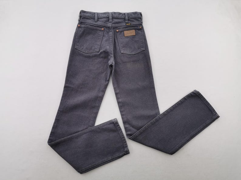 May include: Dark gray denim jeans with a classic five-pocket design. The jeans feature a brown leather patch and copper rivets. The Wrangler brand is visible on the patch. The jeans are laid flat on a white surface.