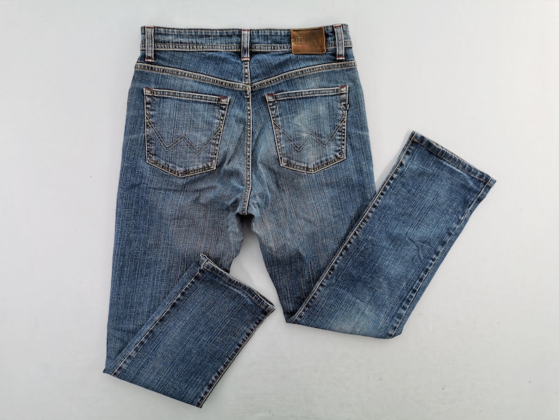 May include: Blue denim jeans with a classic five-pocket design. The jeans have a faded wash, a brown leather patch on the back, and a straight leg cut. The back pockets have a decorative stitch.