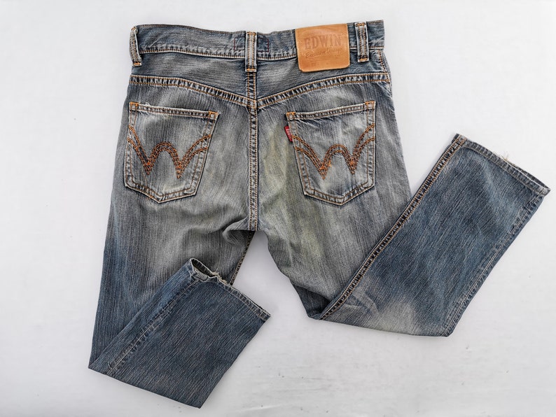 Puede incluir: Vaqueros de mezclilla azul descolorido con un dise&ntilde;o cl&aacute;sico de cinco bolsillos. Cuenta con costuras decorativas en los bolsillos traseros y un parche de cuero con la marca "EDWIN". Los vaqueros tienen un aspecto desgastado y vintage.