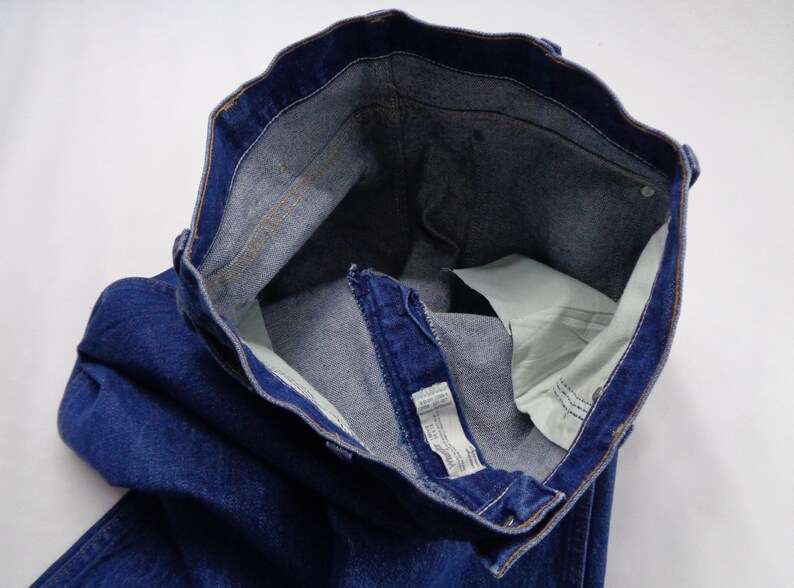 May include: Close-up of a pair of blue denim jeans. The jeans are unbuttoned and unzipped, revealing the interior pockets and waistband. The denim is a deep indigo color, with visible stitching and a light-colored interior.