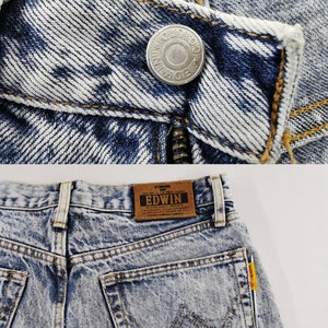 K&ouml;nnte beinhalten: Nahaufnahme einer Jeans aus Acid-Wash-Denim. Die Jeans hat einen silbernen Knopf mit gepr&auml;gtem Text und einen Rei&szlig;verschluss. Auf der R&uuml;ckseite der Jeans befindet sich ein braunes Lederetikett mit der Marke "EDWIN" und eine Tasche mit einem markanten Nahtdesign.