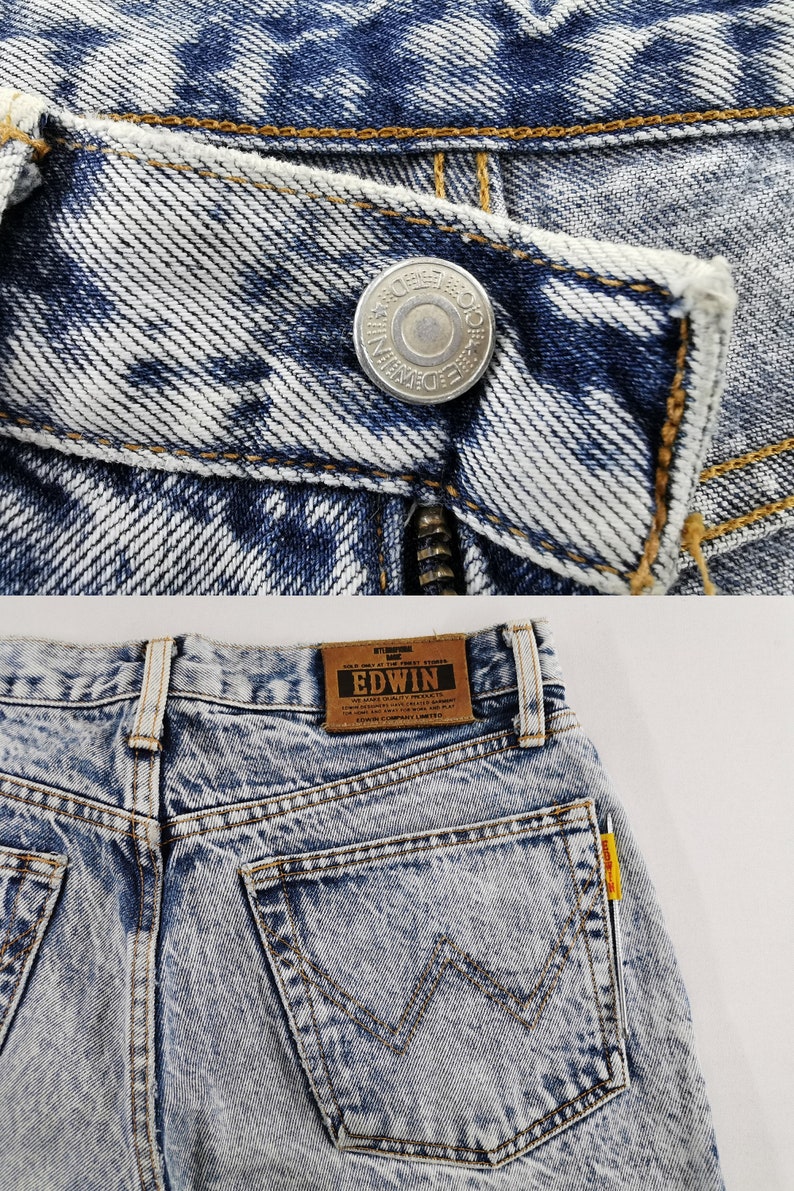 K&ouml;nnte beinhalten: Nahaufnahme einer Jeans aus Acid-Wash-Denim. Die Jeans hat einen silbernen Knopf mit gepr&auml;gtem Text und einen Rei&szlig;verschluss. Auf der R&uuml;ckseite der Jeans befindet sich ein braunes Lederetikett mit der Marke "EDWIN" und eine Tasche mit einem markanten Nahtdesign.