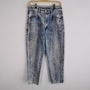 May include: Acid wash denim jeans with a high waist and straight leg. The jeans have a light blue and gray marbled pattern, with vertical seams down the front and back of the legs. They are hanging on a wooden hanger.