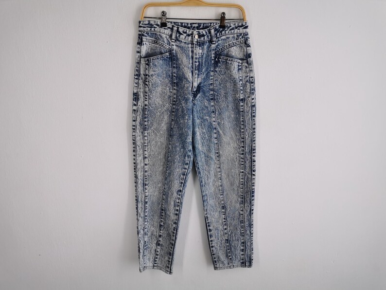 May include: Acid wash denim jeans with a high waist and straight leg. The jeans have a light blue and gray marbled pattern, with vertical seams down the front and back of the legs. They are hanging on a wooden hanger.