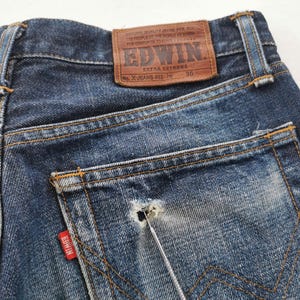 K&ouml;nnte beinhalten: Nahaufnahme der R&uuml;ckseite einer blauen Jeans. Die Jeans hat einen braunen Lederaufn&auml;her mit dem Markennamen "EDWIN" und "EXTRA EXTREME". Ein rotes Etikett mit dem Markennamen befindet sich auf der Ges&auml;&szlig;tasche. Die Jeans hat einen Used-Look mit einem Loch in der Tasche.