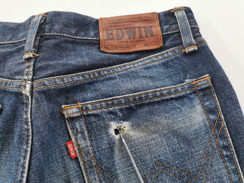 K&ouml;nnte beinhalten: Nahaufnahme der R&uuml;ckseite einer blauen Jeans. Die Jeans hat einen braunen Lederaufn&auml;her mit dem Markennamen "EDWIN" und "EXTRA EXTREME". Ein rotes Etikett mit dem Markennamen befindet sich auf der Ges&auml;&szlig;tasche. Die Jeans hat einen Used-Look mit einem Loch in der Tasche.