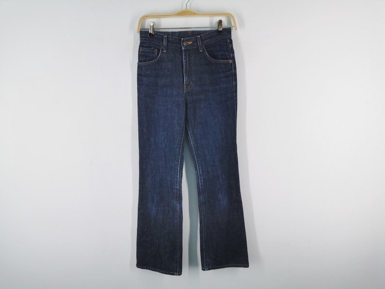 May include: Dark wash denim jeans with a high waist and flared leg, hanging on a wooden hanger. The jeans have classic five-pocket styling and a button and zipper closure. The fabric appears to be a medium weight.