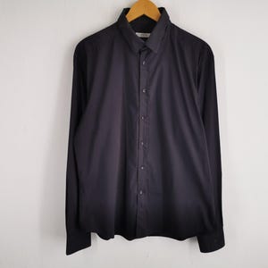 May include: A black long-sleeve button-down shirt hanging on a wooden hanger. The shirt has a classic collar and buttoned cuffs. The shirt is made of a smooth, solid-colored fabric.