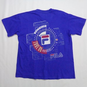 May include: A royal blue t-shirt with a graphic design on the back. The design includes the FILA logo, text that reads "OBJECT OF THIS GAME", "TAKE NO PRISONERS", and other text. The shirt is short-sleeved.