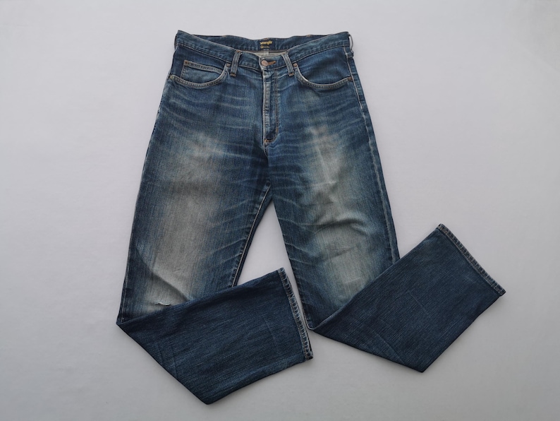 May include: Blue denim jeans with a faded wash and a classic five-pocket design. The jeans have a button and zipper closure and are laid flat on a white surface. The jeans are a medium wash with some fading.