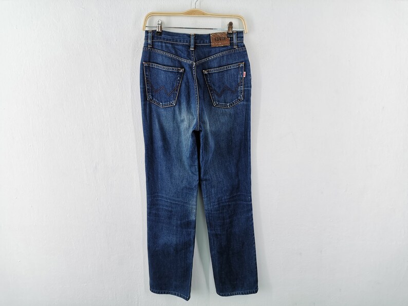 May include: Dark wash denim jeans hanging on a wooden hanger. The jeans have a high waist, straight leg, and classic five-pocket design. A brown leather patch with the brand name is visible at the back.
