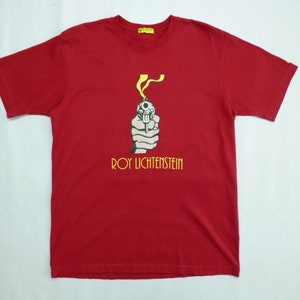 May include: Red t-shirt featuring a graphic of a hand holding a gun with yellow smoke and the text "ROY LICHTENSTEIN" in yellow. The shirt is a solid red color.