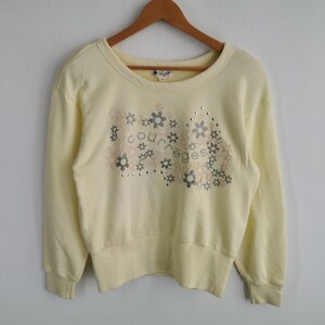 May include: Pale yellow Courreges sweatshirt with a crew neck and long sleeves. The front features a floral design with the brand name and small silver studs. The sweatshirt is hanging on a wooden hanger.