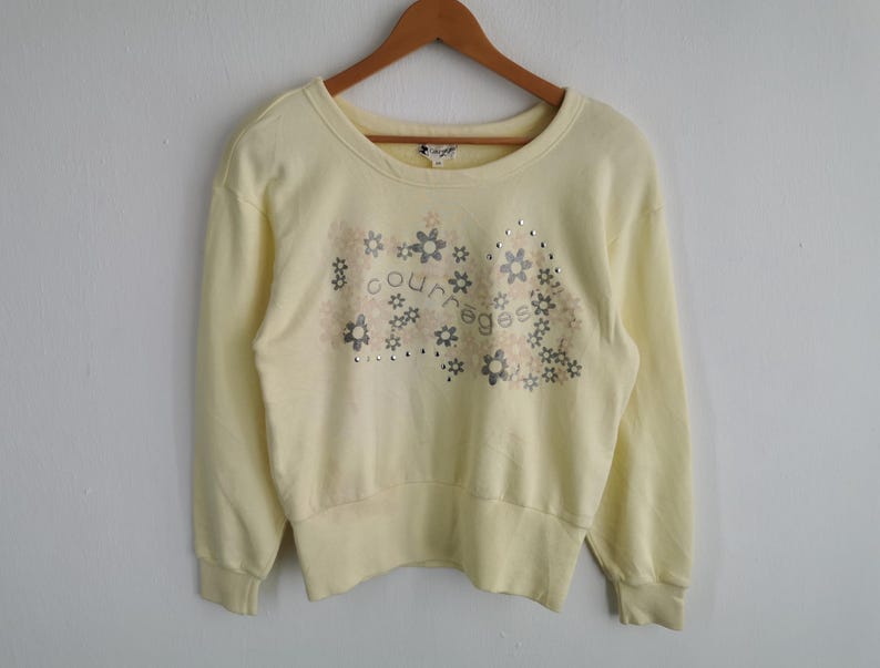 May include: Pale yellow Courreges sweatshirt with a crew neck and long sleeves. The front features a floral design with the brand name and small silver studs. The sweatshirt is hanging on a wooden hanger.