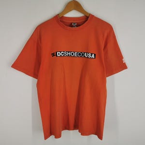 May include: A bright orange short-sleeve t-shirt featuring a crew neckline and a black rectangular graphic on the chest with the white text "DCSHOECOUSA". A small white DC logo is also visible on the left sleeve of the orange cotton garment.