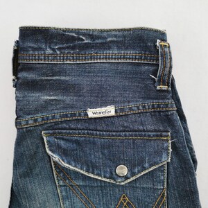 Puede incluir: Primer plano de unos vaqueros de mezclilla azules. Los vaqueros tienen un bolsillo trasero con cierre de bot&oacute;n y el nombre de la marca "Wrangler" en una peque&ntilde;a etiqueta. Las costuras son de color amarillo contrastante.