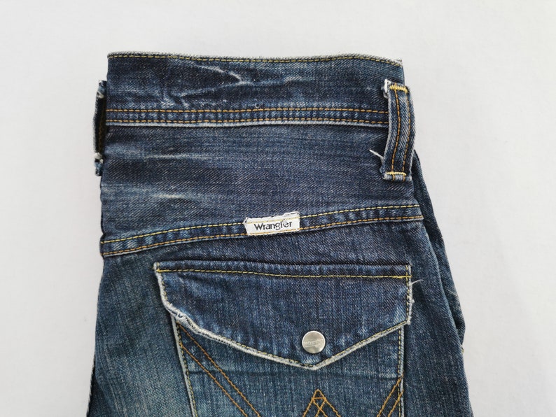 Puede incluir: Primer plano de unos vaqueros de mezclilla azules. Los vaqueros tienen un bolsillo trasero con cierre de bot&oacute;n y el nombre de la marca "Wrangler" en una peque&ntilde;a etiqueta. Las costuras son de color amarillo contrastante.
