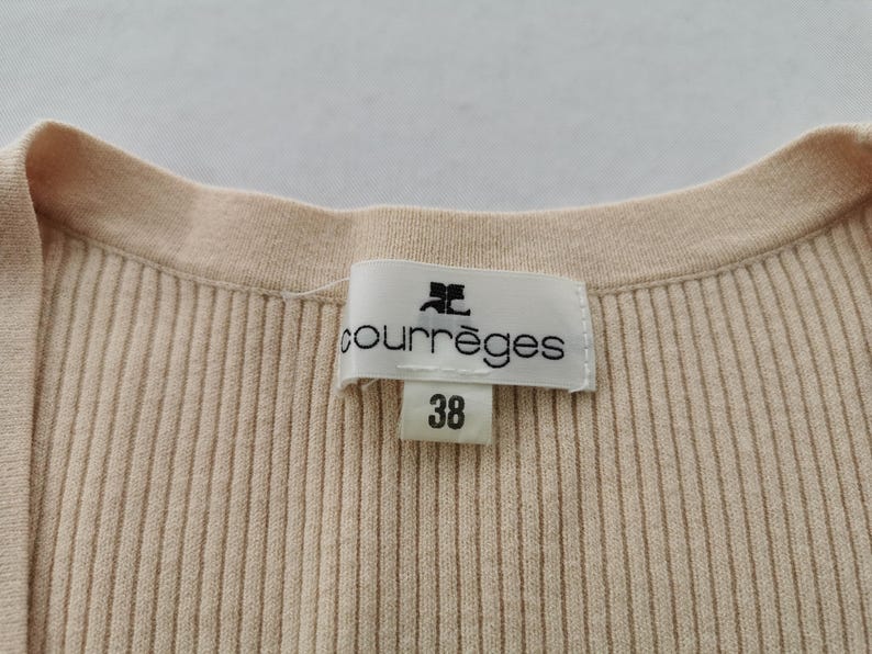 May include: Close-up of a beige ribbed knit top with a round neckline. A label with the brand name "courr&egrave;ges" and the size "38" is visible. The fabric has vertical ribbing.