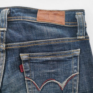 Pu&ograve; includere: Jeans in denim blu scuro con una toppa in pelle marrone sulla cintura, passanti per cintura e una tasca posteriore con un'etichetta rossa del marchio Edwin. La tasca presenta un motivo a onde cucito in grigio e rosso con cuciture arancioni.