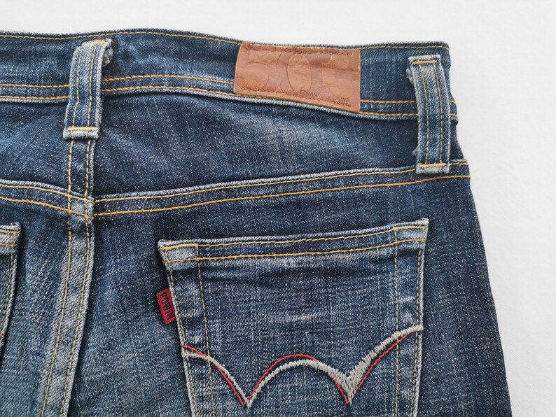 Pu&ograve; includere: Jeans in denim blu scuro con una toppa in pelle marrone sulla cintura, passanti per cintura e una tasca posteriore con un'etichetta rossa del marchio Edwin. La tasca presenta un motivo a onde cucito in grigio e rosso con cuciture arancioni.
