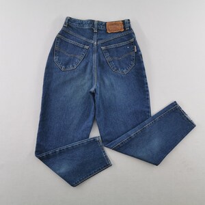 May include: Blue denim jeans with a high waist and straight leg. The jeans have classic five-pocket styling, a button and zipper closure, and a leather patch on the back waistband. The jeans are a medium wash.