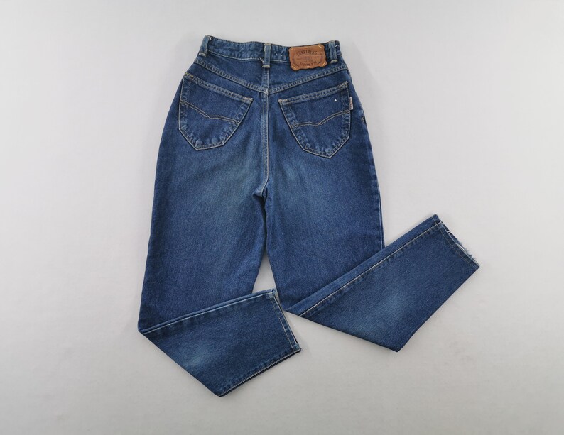 May include: Blue denim jeans with a high waist and straight leg. The jeans have classic five-pocket styling, a button and zipper closure, and a leather patch on the back waistband. The jeans are a medium wash.