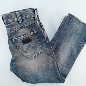 May include: Blue denim jeans with a faded wash. The jeans feature a classic five-pocket design, a black leather patch, and the Wrangler logo on the back pocket. The jeans are folded and laid flat.