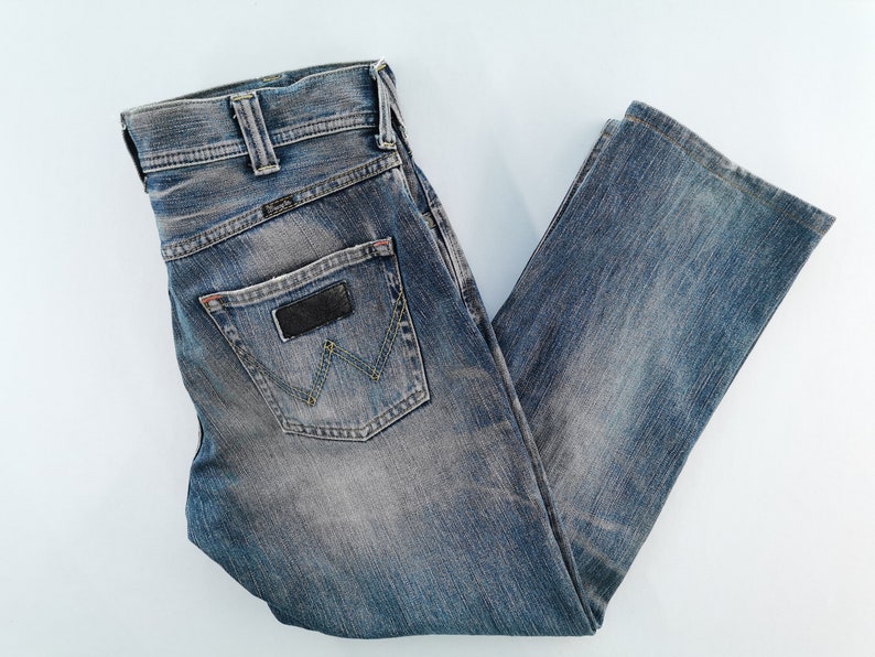 May include: Blue denim jeans with a faded wash. The jeans feature a classic five-pocket design, a black leather patch, and the Wrangler logo on the back pocket. The jeans are folded and laid flat.