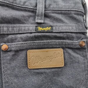 May include: Close-up of gray denim jeans with the Wrangler logo on the back pocket and waistband. The jeans have copper-colored rivets and a brown leather patch with the Wrangler logo.