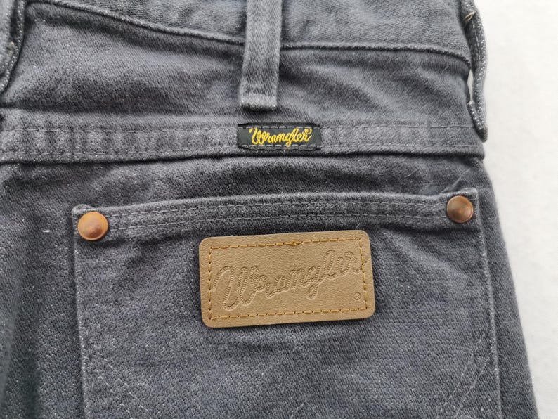 May include: Close-up of gray denim jeans with the Wrangler logo on the back pocket and waistband. The jeans have copper-colored rivets and a brown leather patch with the Wrangler logo.