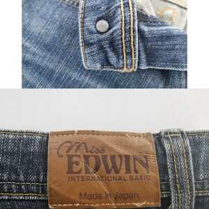 K&ouml;nnte beinhalten: Blaue Jeans aus Denim mit einem Metallknopf, der mit dem Wort EDWIN gepr&auml;gt ist. Ein brauner Lederaufn&auml;her am Bund zeigt den Text Miss EDWIN INTERNATIONAL BASIC und Made in Japan, vern&auml;ht mit kontrastierendem goldfarbenem Garn.